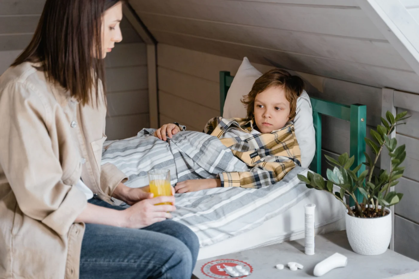 Mother sitting beside her child in bed, offering a warm drink for comfort at home